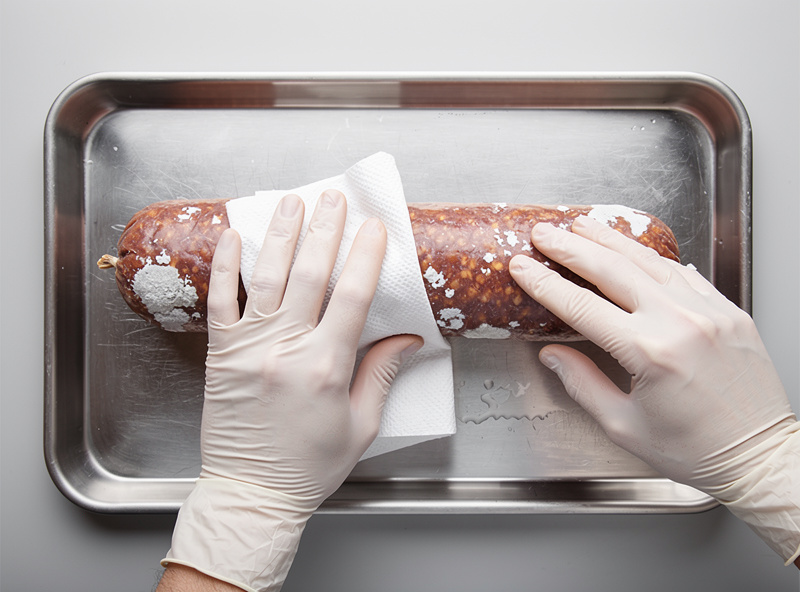 Hands wiping a salami casing with a vinegar-soaked cloth to remove unwanted mold growth over a clean stainless tray
