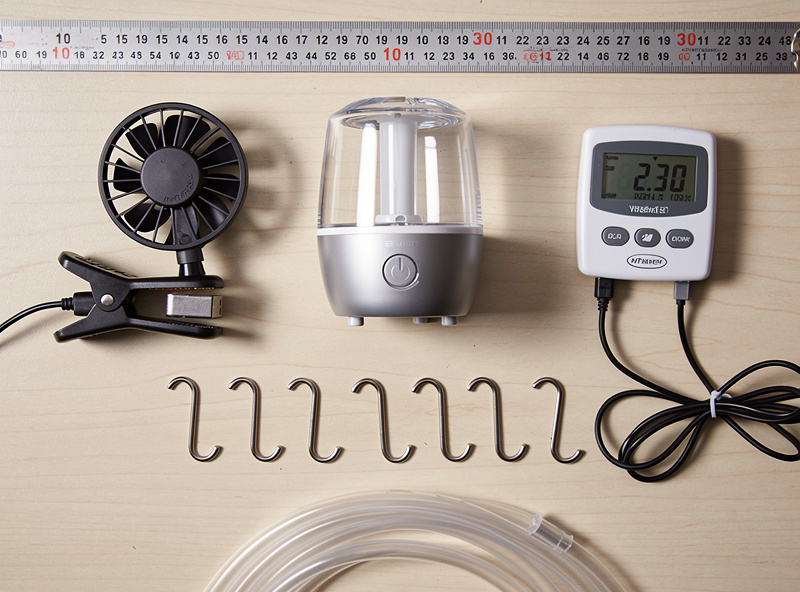 Top-down workbench shot of an ultrasonic humidifier, a small clip-on USB fan, a digital hygrometer probe, and stainless steel hooks