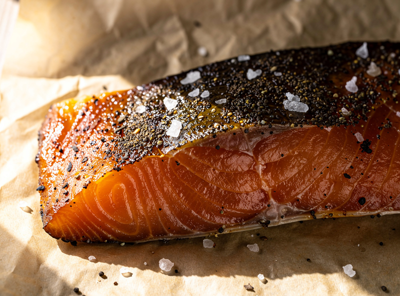 Side of cured salmon (lox) on parchment showing dark amber cold-smoked surface with salt and pepper crust