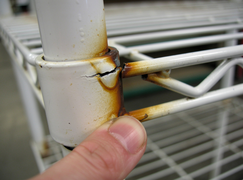 Close-up of cracked white plastic coating on wire shelf with orange rust seeping from the weld joint