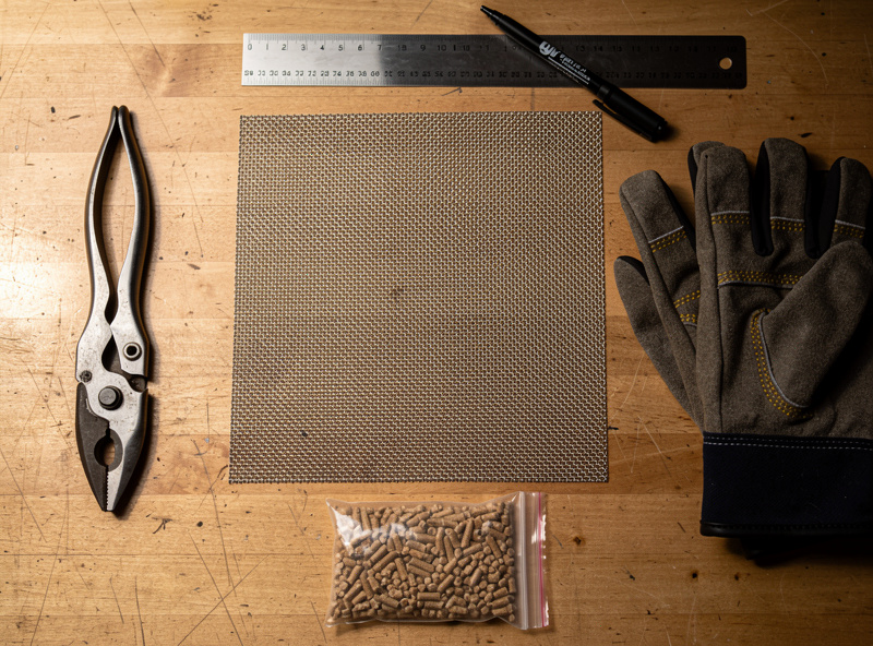 Top-down photograph of materials laid out for a DIY cold smoke generator build — stainless steel mesh sheet, tin snips, ruler, marker, work gloves, and a small bag of food-grade wood pellets