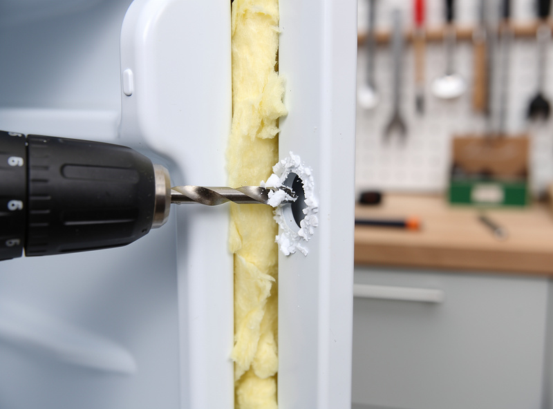 Step drill bit cutting through a mini fridge door from the inside, foam core visible