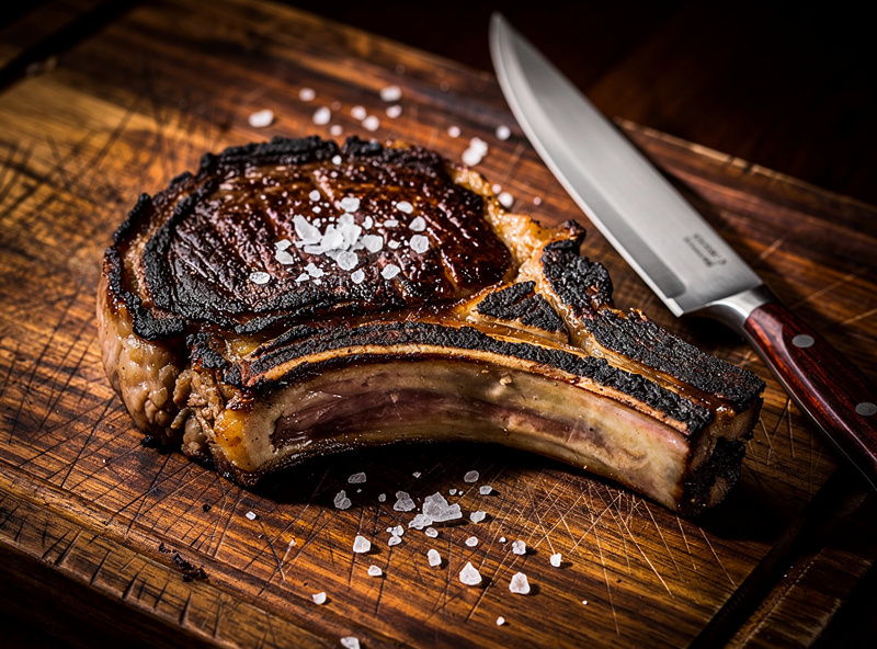 Thick dry-aged ribeye steak seared dark on the outside resting on a wooden cutting board with flaky salt