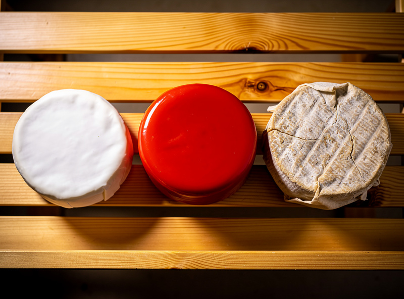 Three wheels of cheese at different ages on a slatted cedar shelf — young white rind, waxed, and cloth-bound natural