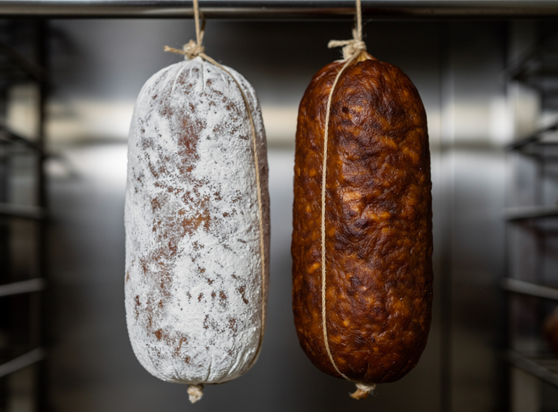 Macro close-up of two cured salamis hanging side by side — one with beautiful white penicillium mold bloom across the surface, the other case-hardened mahogany and shrunken from too-low humidity
