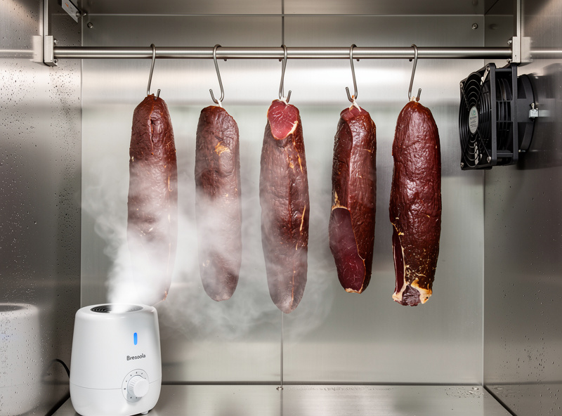 Curing chamber interior with whole-muscle bresaola hanging on stainless steel hooks, humidifier and recirculation fan visible