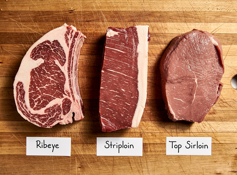 Three beef subprimals labeled ribeye striploin and sirloin laid out on a butcher block for comparison
