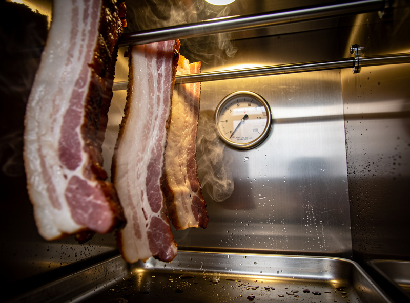 Inside view of stainless smoker with hanging rods, bacon slabs suspended, smoke visible swirling around meat, temperature gauge on wall