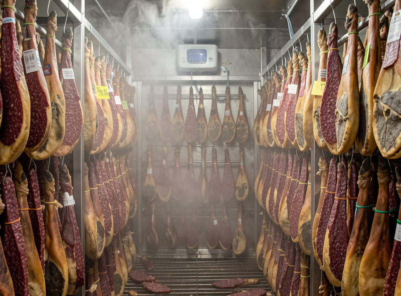 Large curing chamber interior fully loaded with hanging salami and coppa, mist visible from a humidifier