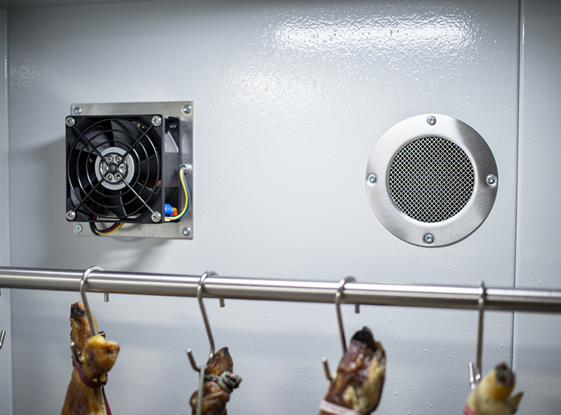 Interior of a curing chamber showing a small back-wall fan and a screened passive vent hole with hanging coppa and salami