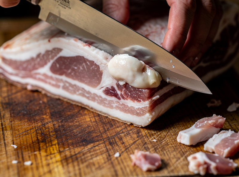 A raw pork jowl skin-on being trimmed on a board, the salivary gland being cut away from the fatty tissue with a boning knife