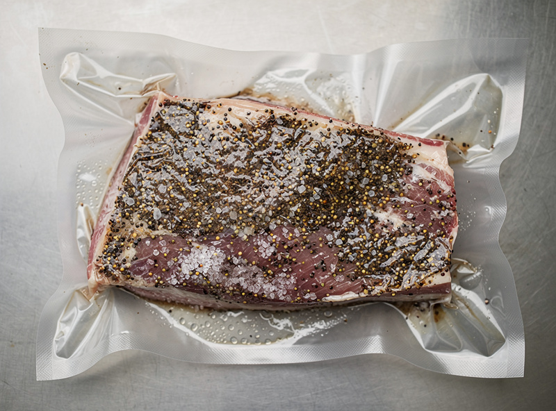 A pork jowl coated in a heavy crust of cracked black pepper and salt cure, sitting in a vacuum bag ready for equilibrium curing