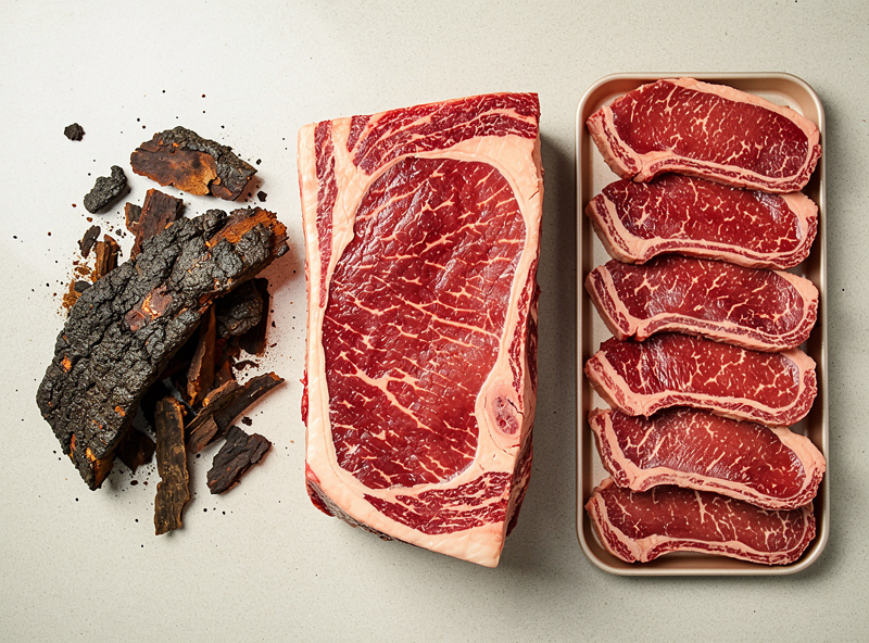 A trimmed dry-aged ribeye subprimal beside a pile of discarded dark bark and a tray of portioned steaks