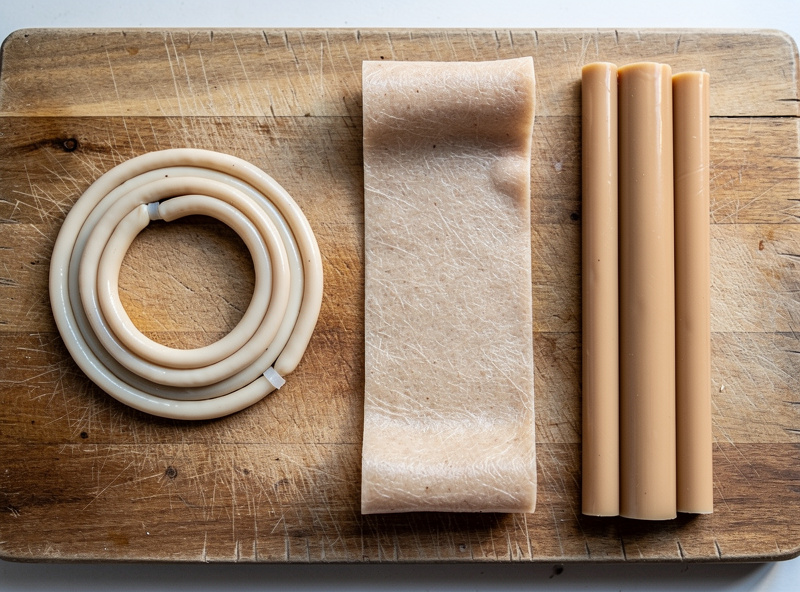 Three types of sausage casing laid side by side on a wooden board: coiled natural hog casing, a wide flat beef middle, and a straight collagen casing tube