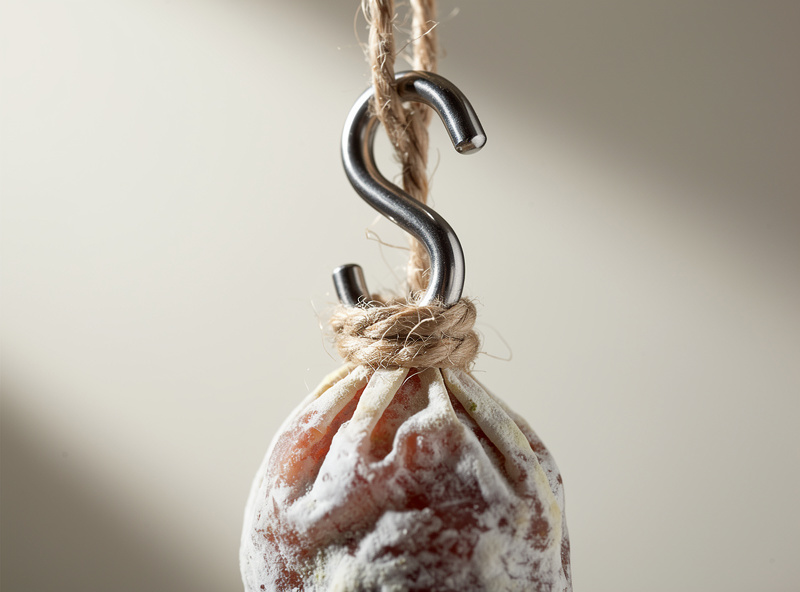 Close-up of a stainless steel S-hook threaded through butcher twine knotted around the tied end of a hanging salami with white mold bloom