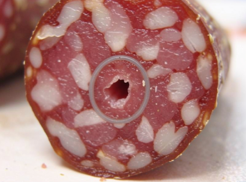 Close-up cross-section of a cut dry salami showing a small trapped air pocket void near the casing with a faint grey ring
